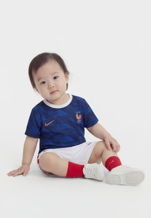 Småbarn sidder på hvidt gulv iført en blå fransk fodboldtrøje, hvide shorts, røde sokker og hvide sko og ser let hældt til siden.