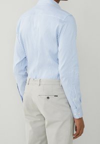 Chemise rayée bleu et blanc avec col boutonné, associée à un pantalon beige clair équipé d'une poche arrière et d'un détail d'étiquette.