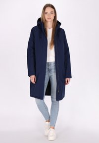 Marineblauwe waterdichte parka met een capuchon, rits- en drukknoopsluiting, lange mouwen en elastische accenten. Gekozen over een witte top en lichte jeans.