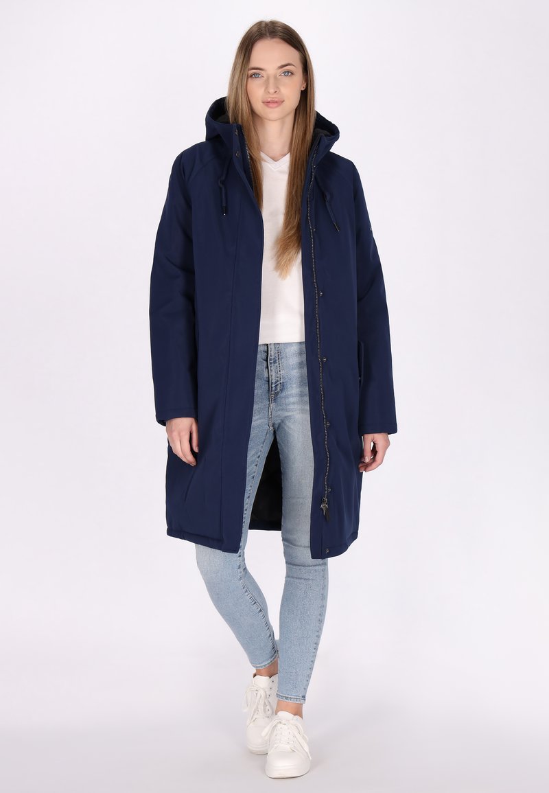 Marineblauwe waterdichte parka met een capuchon, rits- en drukknoopsluiting, lange mouwen en elastische accenten. Gekozen over een witte top en lichte jeans.