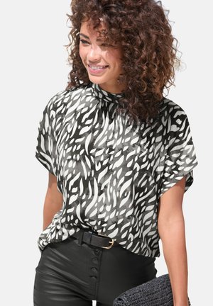 Smiling woman with curly hair wears a black-and-white abstract patterned blouse, black high-waisted pants, and holds a textured black clutch.