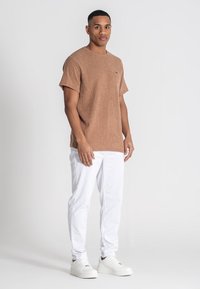 Hnědá texturovaná košile s krátkým rukávem v kombinaci s bílými slim-fit kalhotami a bílými teniskami. Košile má malou nášivku na prsou.