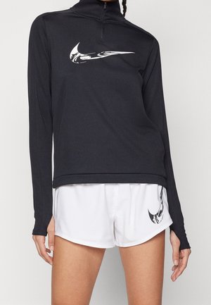 Personne portant un haut à manches longues noir et un short de sport blanc avec des logos Nike swoosh stylisés sur la poitrine et la jambe gauche.
