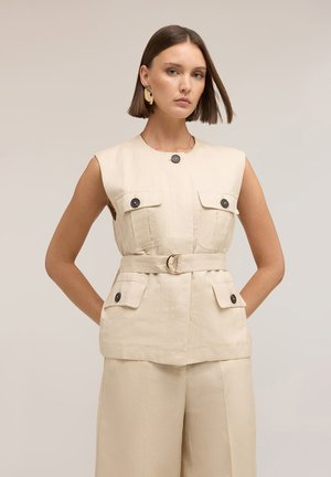 Frau mit kurzem braunem Haar, die eine ärmellose beige Weste mit Gürtel und vier schwarzen Knopftaschen sowie passende weite Hosen vor neutralem Hintergrund trägt.