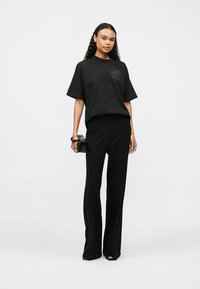 Melna pārspīlēta t-krekla ar nelielu logotipu, kombinēta ar melniem piegulošiem biksēm. Modelis tur mazu rokassomu, stāvot pret vienkāršu fonu.