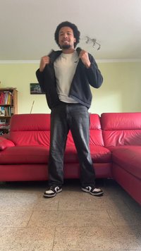 Zwarte fleecejack over een wit T-shirt, donkere jeans en zwart-witte sneakers. Staand in een woonkamer met rode leren banken.