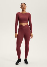 Bordó hosszúujjú crop top és magas derekú leggings sima, rugalmas anyagból. A derékpánton kereszt formájú dizájn található, fekete tornacipővel párosítva.