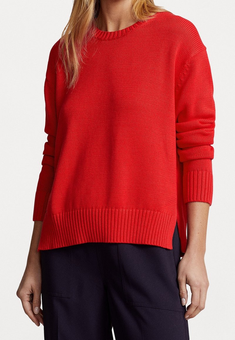 Femme portant un pull en maille rouge vif à col rond avec des poignets et un ourlet côtelés, associé à un pantalon bleu marine foncé sur un fond uni.