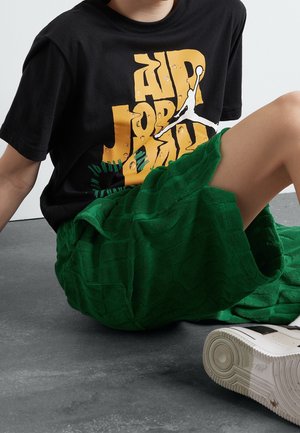 Kind draagt een zwarte Air Jordan T-shirt, groene gestructureerde korte broek en witte sneakers, zittend op een grijze vloer.