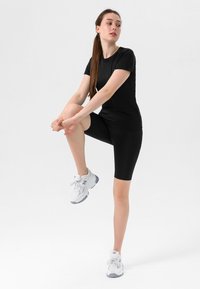 Svart treningsgenser og shorts i et glatt, elastisk materiale. Modell iført hvite og grå joggesko. Minimalistisk bakgrunn.