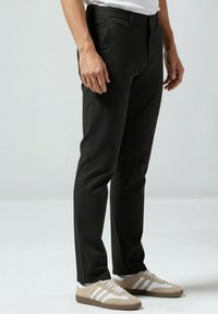 Moški, oblečen v ozke črne hla če in bež superge z belimi črtami, stoji pred enostavnim svetlim ozadjem.