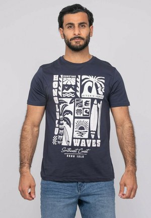 Homme barbu portant un T-shirt bleu marine avec des motifs et textes blancs sur le thème du surf et tropical, associé à un jean bleu, en position neutre.