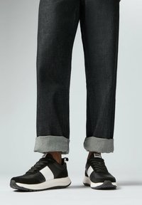 Zwart-witte sportieve sneakers met een textuur bovenkant, voorzien van een witte zijstreep, gedragen met donkere denim jeans, omgeslagen bij de enkels.