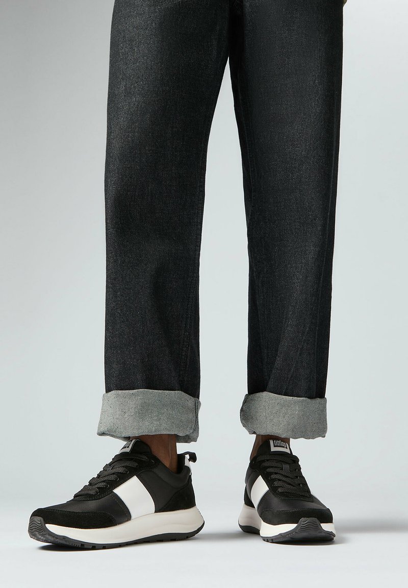 Zwart-witte sportieve sneakers met een textuur bovenkant, voorzien van een witte zijstreep, gedragen met donkere denim jeans, omgeslagen bij de enkels.