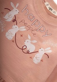 Różowa bluza z czterema białymi królikami z kwiatowymi akcentami, napisem "szczęśliwi ludzie" oraz miękkim, teksturowanym materiałem bawełnianym.