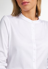 Donna che indossa una camicia bianca con bottoni e colletto alla cinese, mostrando la parte superiore del torso e parte del viso.