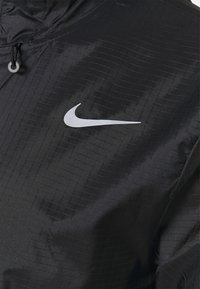 Must mustaga, millel on kerge tekstuuriga kangas. Ees on hõbedane Nike logo ja reguleeritav pael.