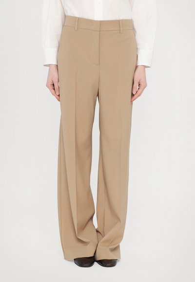 Pantalons beige ajustés avec une coupe droite, taille haute et pinces repassées. Tissu doux à fine texture, associés à des chaussures sombres.