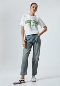 Hvid bomulds t-shirt med grøn og sort grafisk tekst, højtaljede lyse denim jeans og hvide sneakers med sorte accenter.