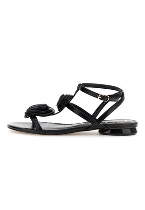 Sandalia negra de tacón bajo con tiras finas, hebilla dorada y detalle superpuesto en forma de pétalos en la tira frontal, suela plana.