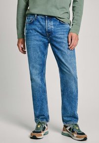 Jeans de mezclilla azul con corte recto, textura desgastada, diseño de cinco bolsillos, combinados con zapatillas deportivas multicolores. Camisa de manga larga verde.