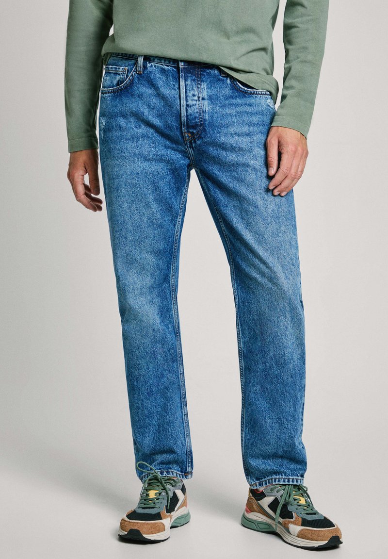 Jeans de mezclilla azul con corte recto, textura desgastada, diseño de cinco bolsillos, combinados con zapatillas deportivas multicolores. Camisa de manga larga verde.