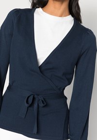 Navy wrap cardigan valmistettu pehmeästä neuloskankaasta, jossa on syvä V-kaula-aukko, pitkät hihat ja solmittava vyö säädettävää istuvuutta varten.