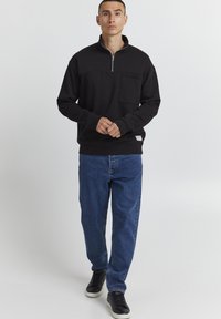 Sort zip-up sweatshirt med høj krave, frontlomme og ribstrikkede manchetter. Kombination med blå jeans og sorte sneakers, stående mod en lys baggrund.