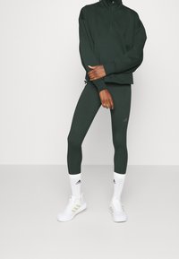Tmavě zelený sportovní outfit skládající se z mikiny na zip a přiléhavých legín, doplněný bílými ponožkami a botami s jemnými akcenty.