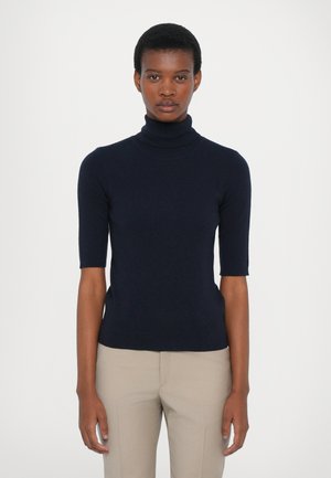 Junge Person mit kurzen Haaren, die ein marineblaues T-Shirt mit kurzen Ärmeln und beige Hosen trägt, steht vor einem schlichten weißen Hintergrund.