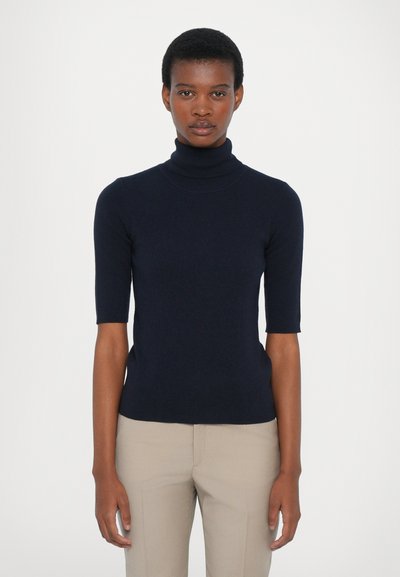 Ung person med kort hår iført en navy blue kortærmet turtleneck og beige bukser, stående foran en ensfarvet hvid baggrund.