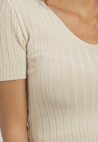 Top en maille de couleur beige clair, avec une texture côtelée, des manches courtes et un décolleté en V profond, complété par une finition en tissu lisse.
