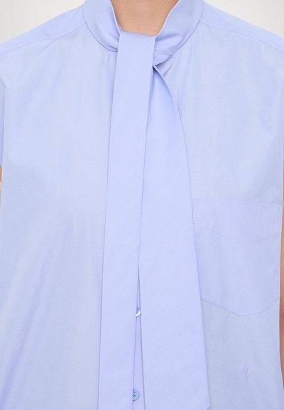 Blusa viola chiaro con colletto a cravatta, maniche corte e tasca a toppa. Il tessuto sembra liscio e leggero, con un leggero riflesso.