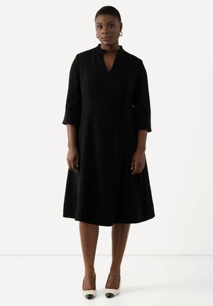 Robe noire midi avec des manches 3/4, un col bateau et une silhouette évasée. Associée à des escarpins à bout pointu bicolores.
