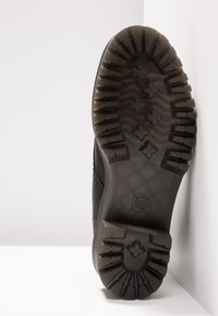 Semelle d'une chaussure noire avec des crampons profonds, des motifs en forme de croix, et un matériau en caoutchouc translucide sur fond blanc.