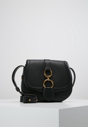 Bolso de cuero negro tipo bandolera con solapa curva, cierre de doble anillo dorado y correa ajustable, sobre pedestal blanco contra fondo gris.