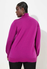 Magenta Pullover mit Stehkragen und langen, puffigem Ärmeln. Weicher Stoff und gerippter Saum, kombiniert mit dunklen Jeans.