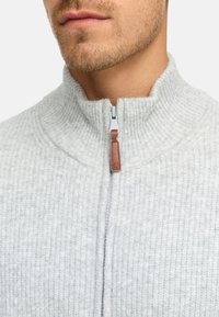Suéter gris claro de canalé con cierre de cremallera y cuello alto, con una cremallera metálica que tiene una tirador de cuero marrón, que muestra una tela texturizada.