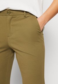 Kalhoty z elastického materiálu v olivově zelené barvě s hladkou texturou, mají zapínání na knoflík, poutka na opasek a boční kapsy. Jednoduchý a funkční design.