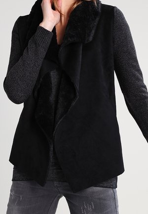 Personne portant un gilet noir sans manches avec un large col doux sur un haut à manches longues foncé et un jean gris délavé sur un fond uni.