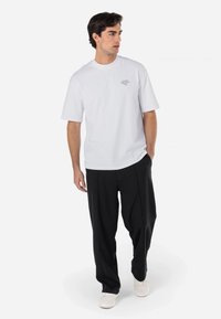 Camiseta de algodón blanca de manga corta, de corte relajado, con un pequeño gráfico púrpura en el pecho. Combinada con pantalones negros holgados y zapatos blancos.