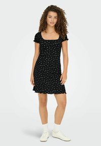 Femme aux cheveux bouclés portant une robe noire à manches courtes avec des pois blancs, des chaussettes blanches et des baskets blanches, se tenant sur un fond uni.