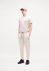 Beige T-Shirt mit kurzen Ärmeln und schwarzem Logo, dazu passende beige Jogginghose und schwarze Sneaker, abgerundet mit einer beige Kappe mit Logo.
