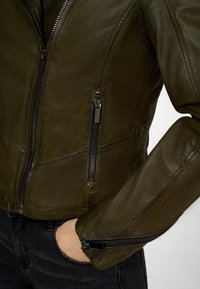 Olivengrøn læderjakke med et tætsiddende design, der har lynlåslommer og en glat overflade. Slanke sorte jeans kan ses.