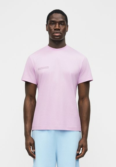 Άνδρας που φοράει ανοιχτό μωβ κοντομάνικο μπλουζάκι με μικρό κείμενο στο στήθος και ανοιχτό μπλε σορτς, στέκεται μπροστά σε ένα απλό λευκό φόντο.