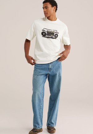 Junger Mann trägt ein weißes T-Shirt mit schwarzer Boombox-Grafik, hellblaue Jeans und braune Schuhe und steht vor einem einfarbigen Hintergrund.