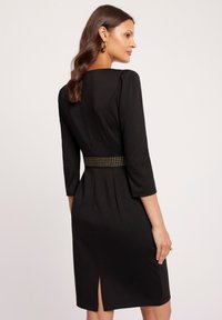 Czarna sukienka z rękawami o długości trzech ćwierć, dopasowanym kroju i przystawką z ćwiekami. Posiada delikatne rozcięcie z tyłu dla dodatkowego akcentu.