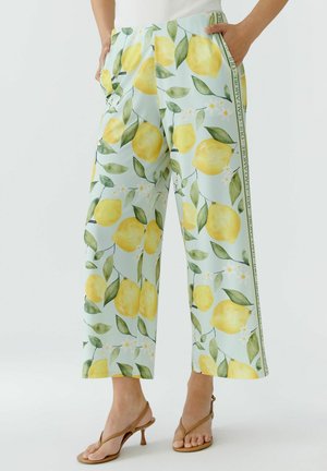 Weite Hose mit gelbem Zitronen- und grünem Blättermuster auf hellblauem Stoff, kombiniert mit braunen Sandalen mit Absatz und einem weißen Oberteil.