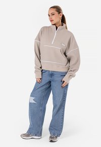 Beige hettegenser med glidelås og teksturert overflate, med hvite detaljer og en frontlomme. Kombinert med lys blå baggy jeans og grå joggesko.