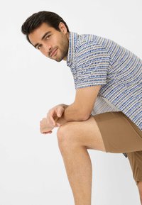 Chemise à manches courtes à boutons avec un motif géométrique bleu et crème. Des shorts marron sont visibles. L'arrière-plan est blanc uni.